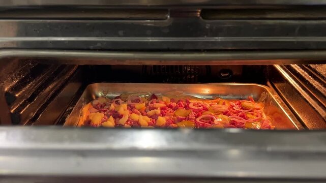 A hand slides a rectangular pizza topped with sausage, chicken, pickles, red onion, and pineapple into an illuminated oven.
