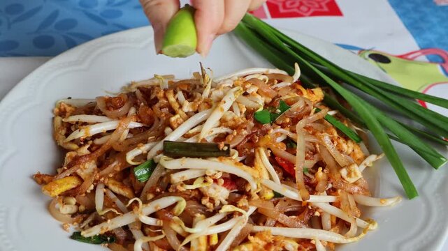 Close up of hands squeezing a fresh juicy lime wedge over a plate of authentic Pad Thai noodles