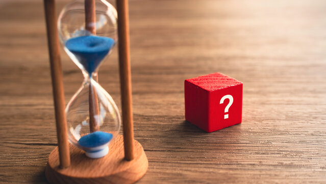 Vertical hourglass with blue sand next to red question mark cube on wooden desk time running out concept