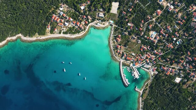 A high-angle top-down aerial view of a picturesque turquoise bay and harbor on Silba Island, Croatia, showing moored boats, lush green vegetation, and vibrant red-roofed houses along the crystal-clear