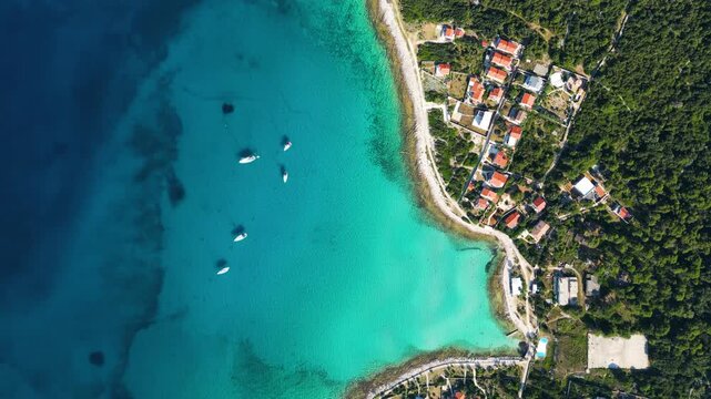 A high-angle top-down aerial view of a picturesque turquoise bay and harbor on Silba Island, Croatia, showing moored boats, lush green vegetation, and vibrant red-roofed houses along the crystal-clear