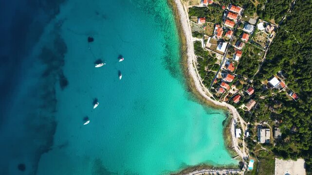 A high-angle top-down aerial view of a picturesque turquoise bay and harbor on Silba Island, Croatia, showing moored boats, lush green vegetation, and vibrant red-roofed houses along the crystal-clear