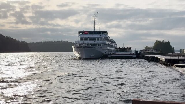 A large cruise ship docks quietly at a pier on sparkling water. It rests peacefully at Lake Ladoga, Karelia under a cloudy sky. The quiet view inspires a sense of peaceful joy.