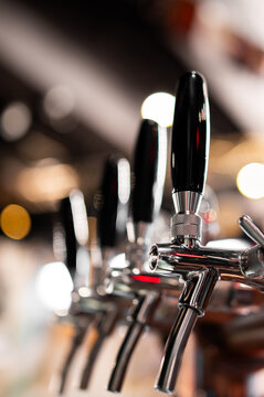 The image shows a close-up of a rows of shiny metal beer taps with black handles at a bar or pub.