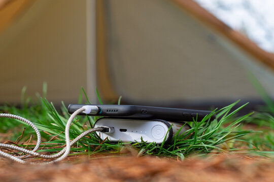 Smartphone charging from portable power bank lying on green grass near camping tent outdoors. Mobile energy supply, outdoor technology, camping gear power source and adventure travel accessory.
