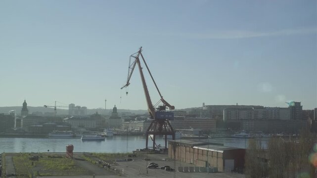 Gothenburg, Sweden - May 2 2023: A large industrial harbor crane stands over the river, with boats moored along the waterfront and the historic city skyline visible in hazy morning light