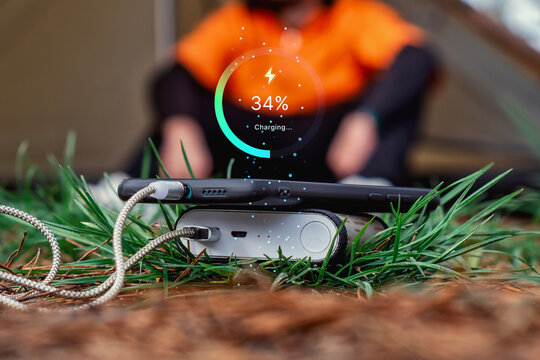 Smartphone connected to a portable power bank on grass with a digital 34 percent charging indicator. Mobile charging, battery power and outdoor technology concept with a person in the background.