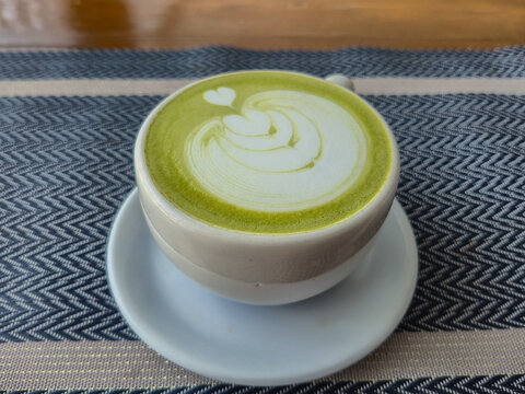 Matcha Green Tea Latte with Heart Foam Art in Ceramic Mug, Top View Flat Lay Angle on Patterned Fabric Tablecloth