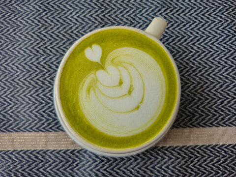 Matcha Green Tea Latte with Heart Foam Art in Ceramic Mug, Top View Flat Lay Angle on Patterned Fabric Tablecloth
