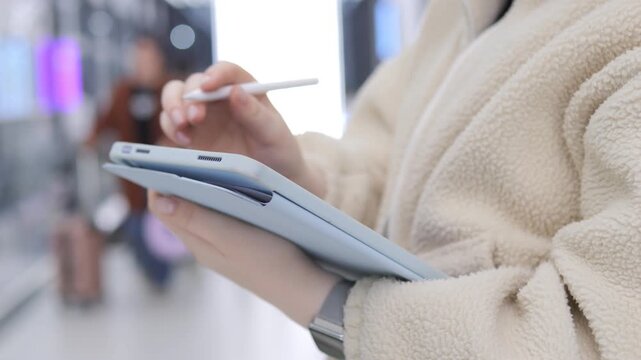 Person in beige fleece jacket holds tablet with stylus. They write or draw on screen while standing. Background shows blurred people walking. Focus stays on hands and device