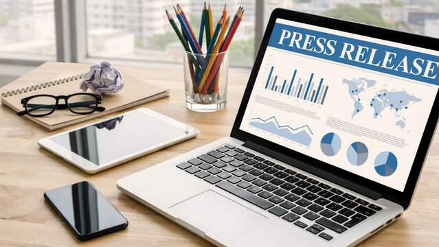 Laptop screen displaying press release data with charts and graphs on a wooden desk with office supplies during daylight