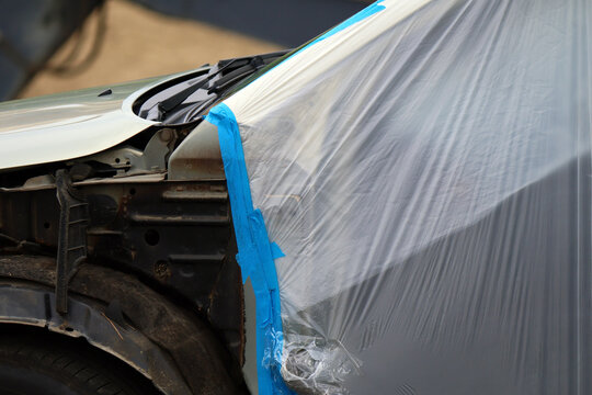 Vehicle with front-end damage features plastic sheeting secured by blue tape to shield undamaged areas; industrial mood highlights auto body repair workflow
