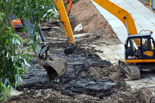 Two yellow tracked excavators operate on a muddy construction site, digging into dark, loose soil beside a paved walkway