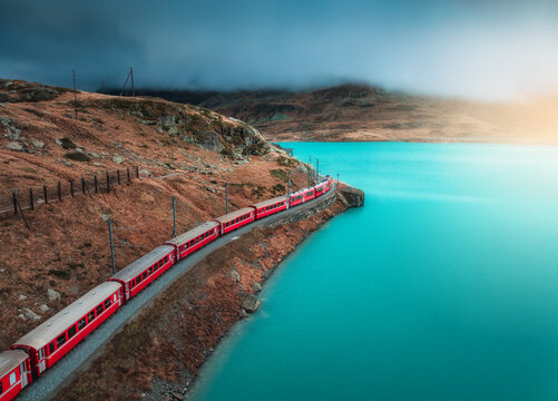 Aerial view of red train moving along turquoise lake in the Swiss Alps, rocky hills, dramatic cloudy sky in autumn alpine mountains. Top view of train in fall. Bernina Express, Switzerland. Railway