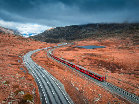 Aerial view of modern red train in alpine mountains, road, orange forest, sky with clouds at sunset in autumn. Bernina Express, Switzerland. Top drone view of train in Alps, winding railroad in fall