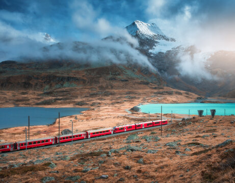 Aerial view of red Bernina Express train, curving railroad, turquoise lake in the Swiss Alps, rocky hills and dramatic cloudy sky at sunset in autumn. Alpine mountains. Top view of train in fall