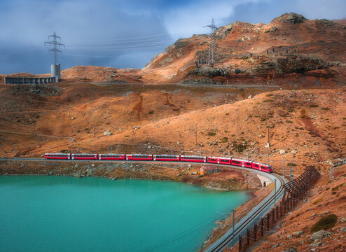 Aerial view of modern red train near azure mountain lake and dramatic cloudy sky at sunset in autumn. Bernina Express, Switzerland. Top view of train in Alps, winding railroad, orange hills in fall