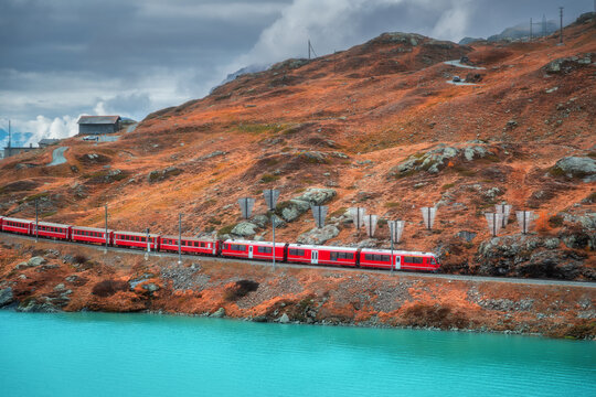 Aerial view of red train moving along the turquoise lake in the Swiss Alps, rocky hills, dramatic cloudy sky in alpine mountains in autumn. Top view of train in fall. Bernina Express, Switzerland