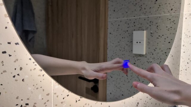 White female hand touching a sensor touch button on a modern round mirror in a contemporary bathroom and turns the integrated LED backlight on and off. The mirror reflects a door and a hanging towel