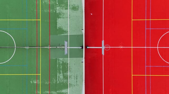 Straight-down drone view of adjacent red and green multisport courts with basketball hoops and overlapping colored markings forming a bold geometric split.
