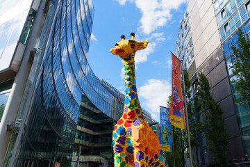 Fototapeta premium Berlin, Germany - August 29, 2026: Low angle view of giant colorful brick giraffe statue framed by curved glass facade reflecting blue sky and clouds in Potsdamer Platz area, Berlin, Germany.