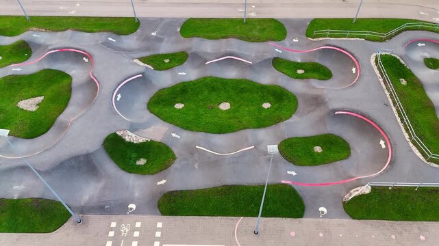 Oblique drone view of an empty pump track with sweeping berms, arrow-marked asphalt lanes and grassy islands forming a complex cycling skills course.