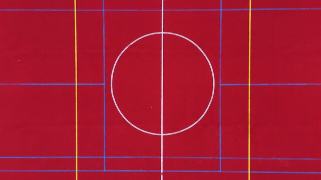 Straight-down drone view of a full red multisport basketball area with four hoops, center circle and layered white, yellow and blue markings forming a striking symmetrical pattern.