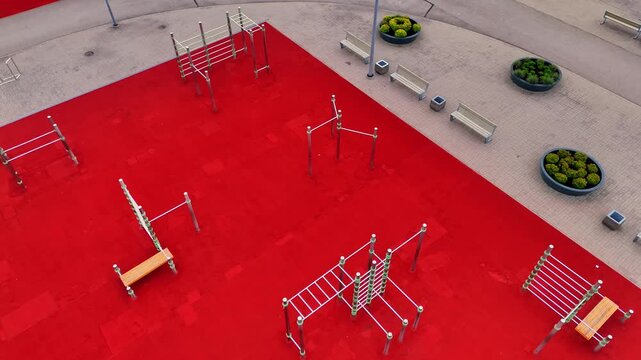 Oblique drone view of an empty calisthenics park with monkey bars, climbing frames and bodyweight stations on vivid red rubber flooring beside benches and planters.