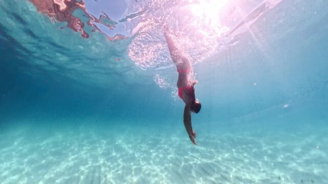Freediving woman in vibrant red bikini gracefully dances underwater with white fins in tropical sea. Wide angle underwater tracking shot.