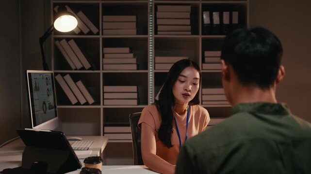 Over the shoulder view of a young asian businesswoman explaining a new business strategy to a male colleague while working late in a modern office, showing dedication and commitment