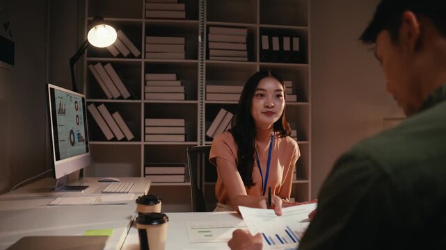 Young Asian business woman presenting financial data on a paper document to her male colleague during a late night meeting in a modern office, showing teamwork and commitment to their project