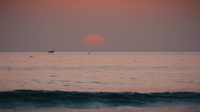 The sun sets over the Atlantic Ocean at Tamraght, Morocco, with a fishing boat and bird silhouetted against the glowing sky.