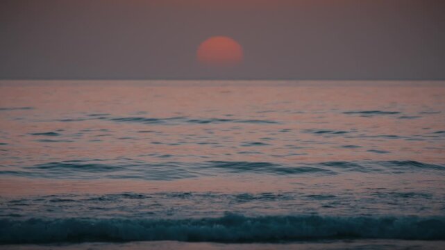 The sun sets over gentle waves on the Atlantic Ocean at Tamraght, Morocco, casting a soft orange glow across the water.