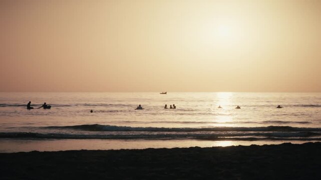 Surfers sit in the ocean at sunset in Tamraght, Morocco, with a fishing boat silhouetted on the glowing horizon.