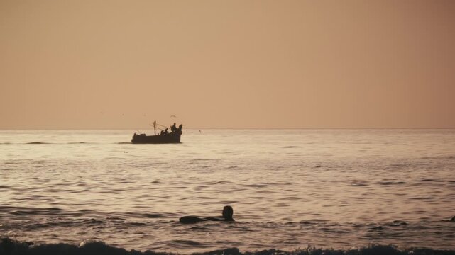 A fishing boat passes as a lone surfer sits in the ocean at sunset in Tamraght, Morocco, with birds over the golden-lit water.
