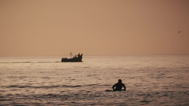 A fishing boat sails past two surfers in the ocean at sunset in Tamraght, Morocco, as birds fly overhead.