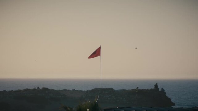 The Moroccan flag waves atop a flagpole above the coastline at sunset in Tamraght, Morocco.
