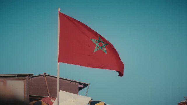 The Moroccan flag waves atop a flagpole above a rooftop against a clear blue sky in Tamraght, Morocco.