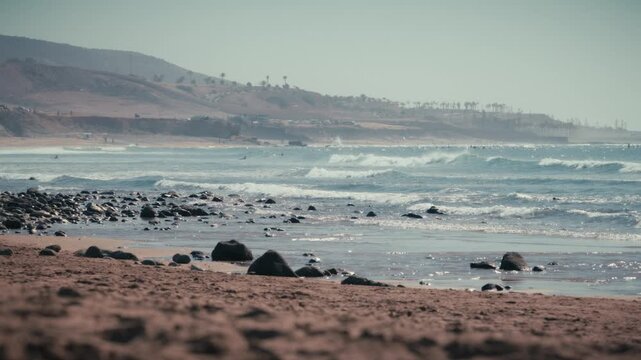 Waves break along the rocky shore of Banana Beach, Tamraght, Morocco, with a palm-lined hillside stretching along the coast behind.