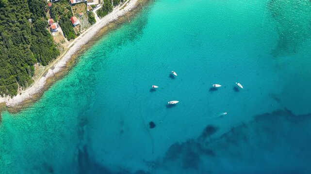 An aerial panoramic view of the tranquil turquoise bay on Silba Island, Croatia, featuring anchored sailboats along a lush, green Mediterranean coastline with charming red-roofed village houses under