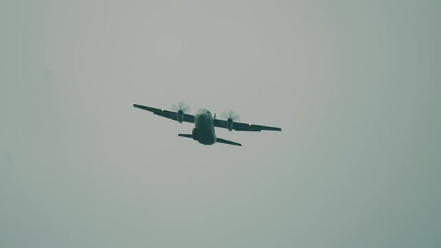 Twin engine military cargo airplane flies directly overhead in gray overcast skies during tactical Falcon Leap exercise in Oosterbeek Netherlands filmed from below.