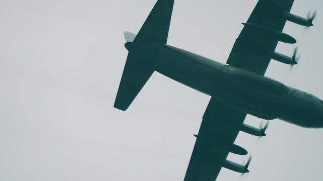 Military transport airplane performs low flyby overhead against overcast skies during Falcon Leap commemoration event in Oosterbeek Netherlands in tracking shot.