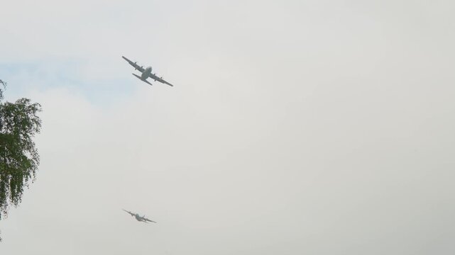 Wide view tracking military turboprop aircraft soaring across cloudy sky past treetop foliage during Operation Market Garden airborne commemoration maneuver.