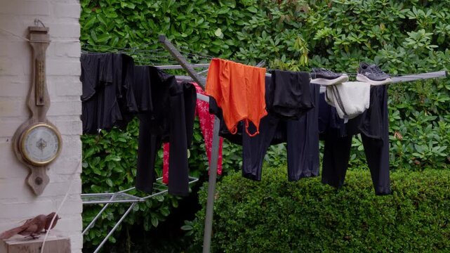 Static wide shot of wet laundry and sneakers hanging on outdoor rotary clothesline next to brick patio wall and green garden hedge during rainy morning in Budel Netherlands.
