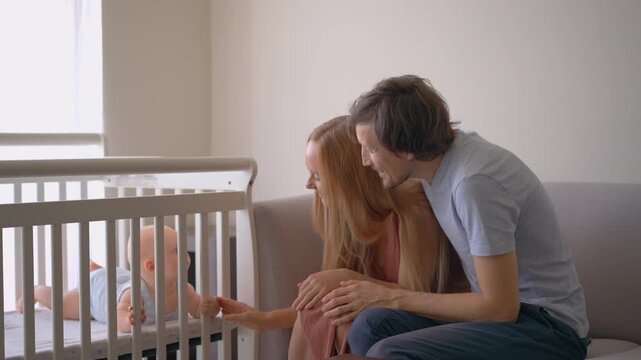 Loving parents watching baby lying in crib, smiling and talking to him.