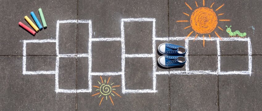 A hopscotch game drawn with chalk on pavement, complete with colorful chalk sticks, two shoes, and whimsical sun and caterpillar drawings, evokes childhood joy and outdoor play.