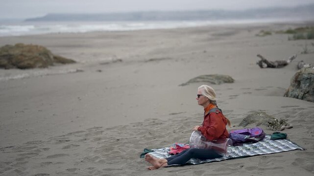 A senior woman plays a doumbek drum while sitting on the beach in Fort Bragg, Mendocino, California, enjoying the coastal environment.