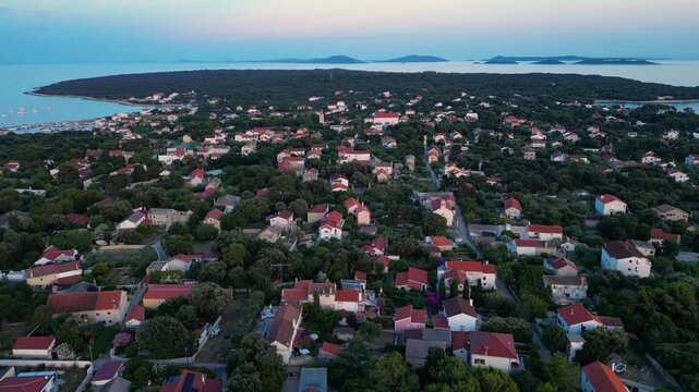 A breathtaking evening aerial drone panorama of old village on Silba Island, Croatia. The shot showcases the vibrant turquoise harbor, a docked ferry, and the charming coastal village stretching