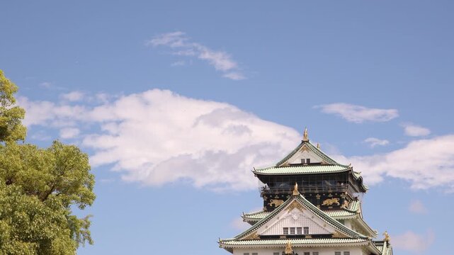 View of the iconic main tower of Osaka Castle in Japan, rising above surrounding trees and showcasing traditional Japanese fortress architecture under a bright sky.