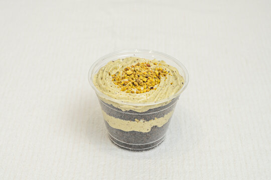 Layered pistachio cream cup dessert with cookie crumble base, piped swirl top, and chopped pistachio garnish on linen backdrop.